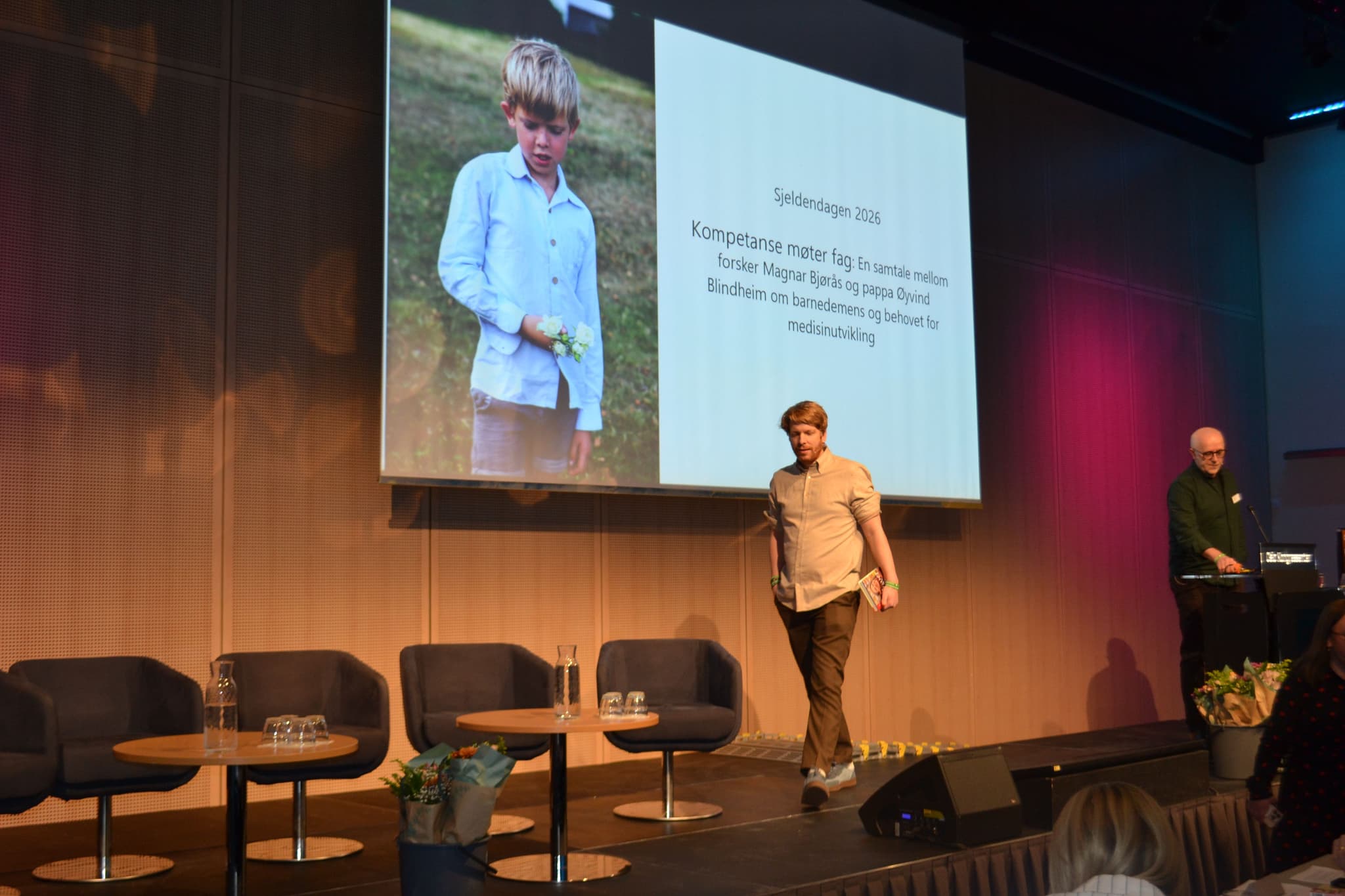 To personer står på en scene. De har på seg skjorter og dressbukse.