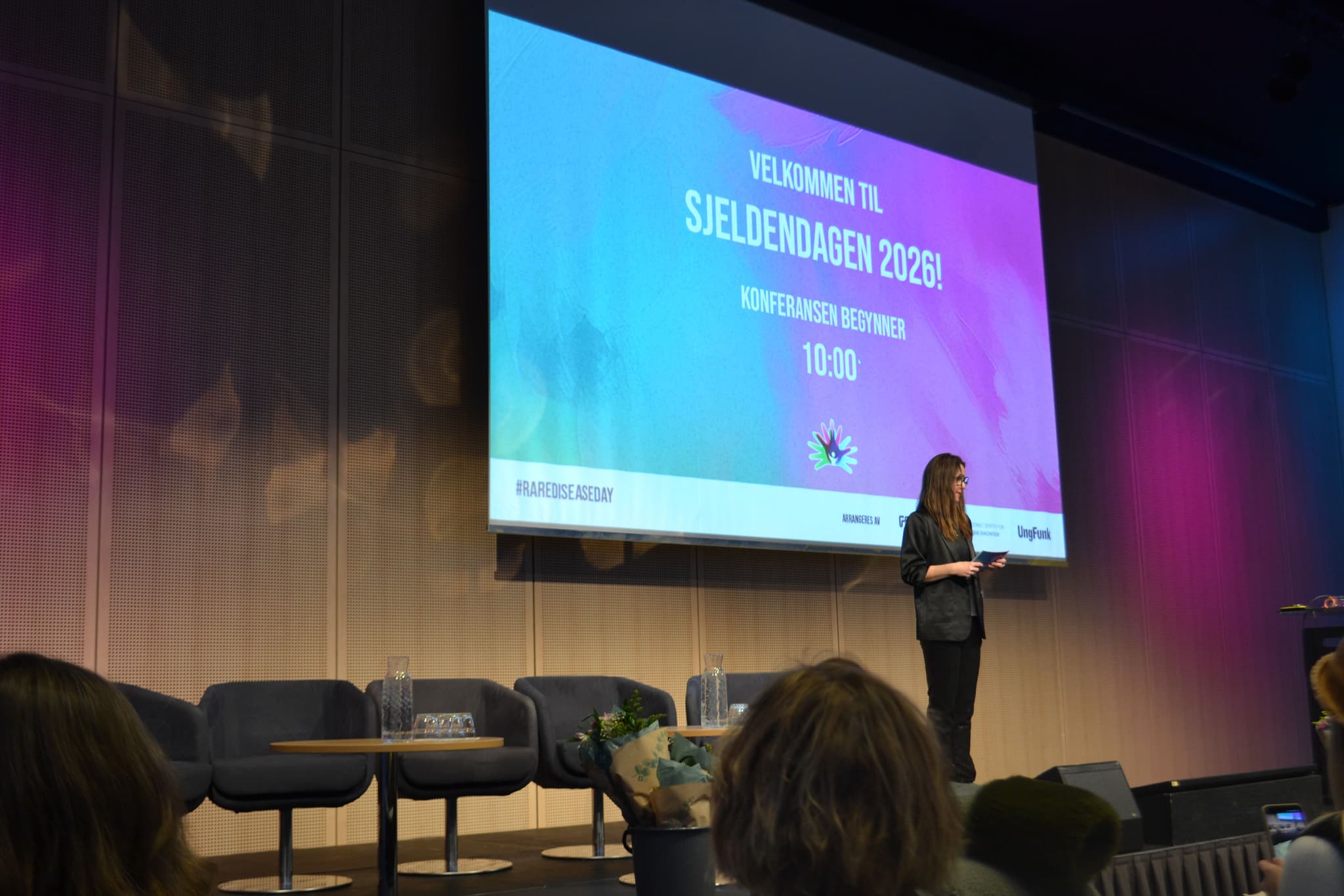 En person står på en scene og presenterer. Hun har på seg dress. Bak hun står det en presentasjon som heter Sjeldendagen 2026.