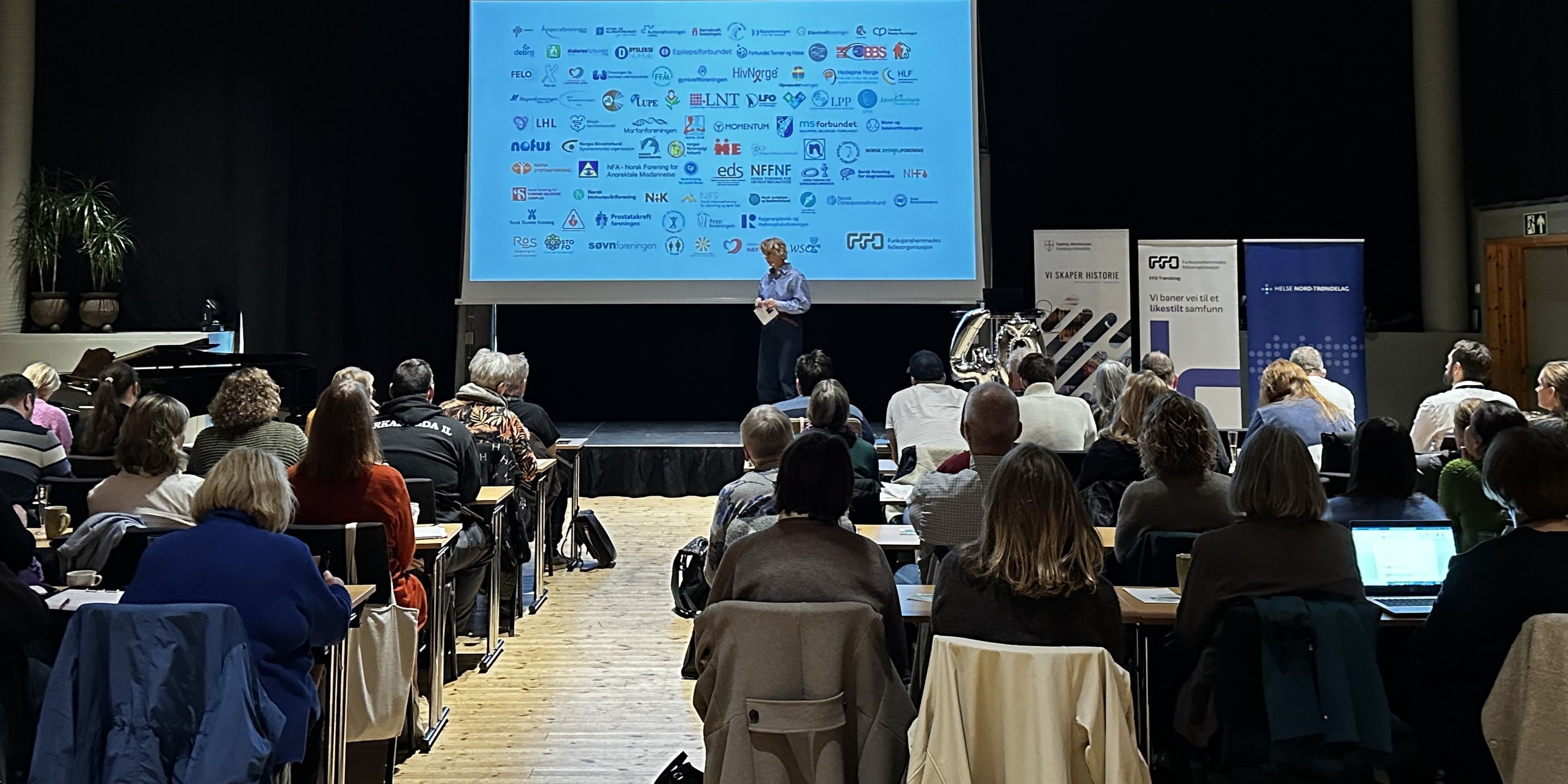 Bilde av møteforsamling sett bakfra. På skjermen er det bilde av logoen til alle medlemsorganisasjonene i FFO.